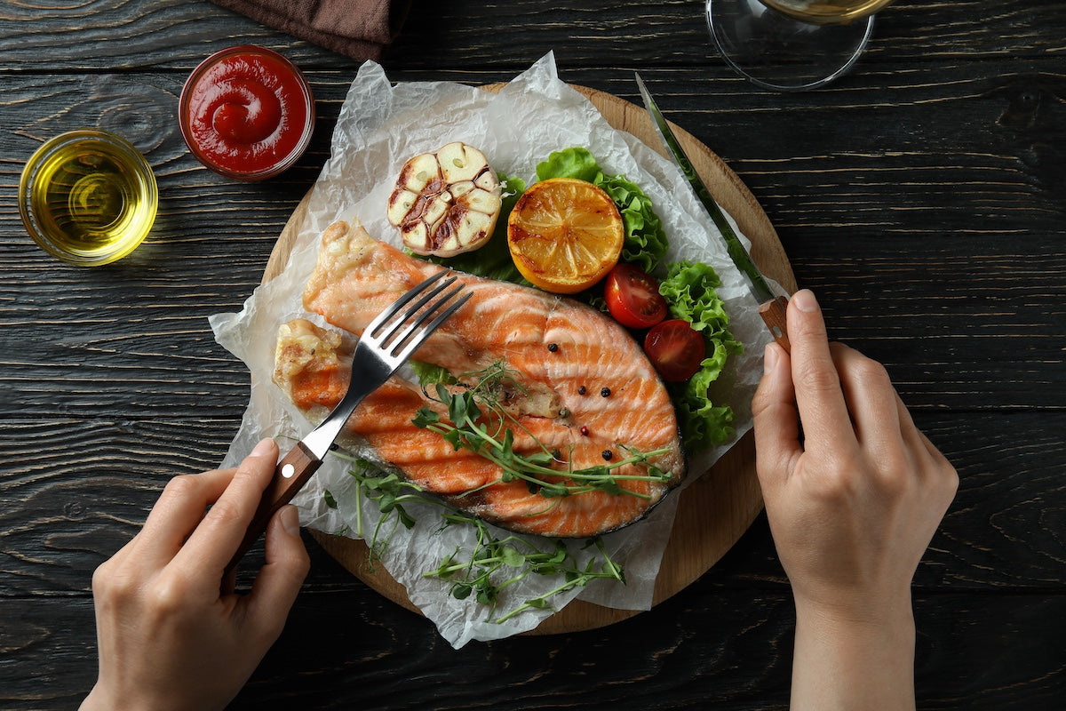 Top view of hands holding fork and knife over a grilled salmon steak served with roasted garlic, lemon, cherry tomatoes, and leafy greens on parchment paper, with olive oil and ketchup on the side.