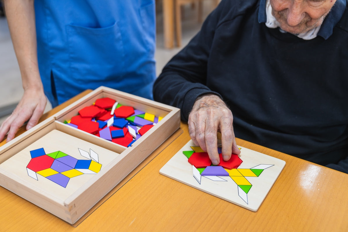 Elderly man solving geometric puzzle for cognitive stimulation with caregiver support — brain training activity to prevent memory decline and Alzheimer’s progression