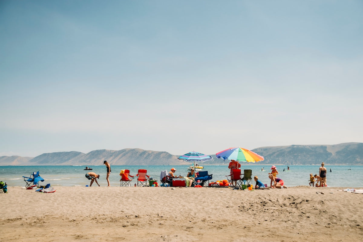 Families relaxing and playing under colorful umbrellas on a sandy beach with clear blue water and mountains in the background on a sunny summer day.