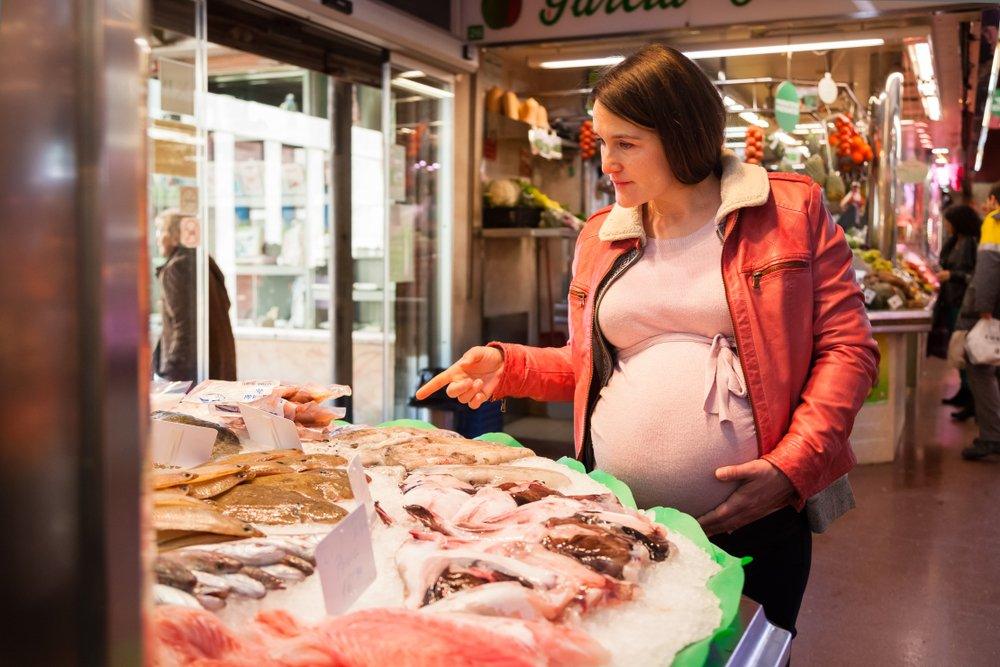 Pregnant woman shopping at a fish market, examining various types of fresh seafood on display, while placing a hand on her belly.