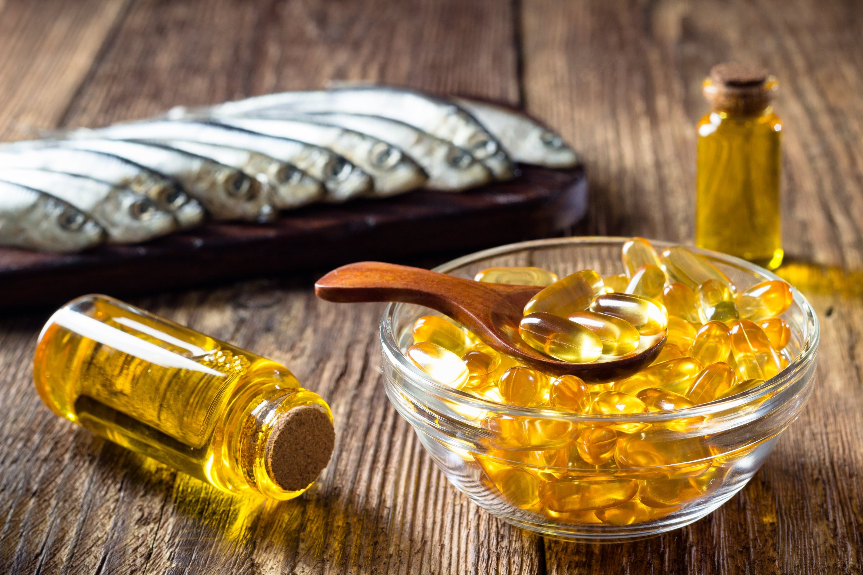 A bowl of golden fish oil capsules with a wooden spoon, two glass bottles of fish oil, and a tray of fresh sardines in the background on a rustic wooden surface.