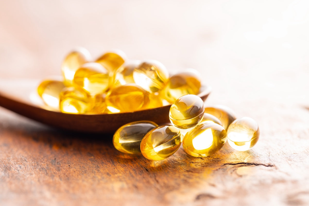 Close-up of golden fish oil capsules on a wooden spoon and rustic wooden surface, illuminated by warm light.