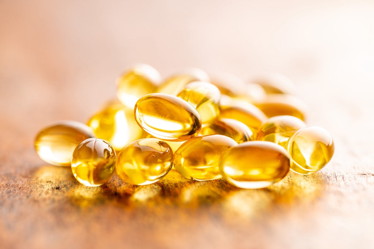 A close-up image of translucent yellow omega-3 fish oil capsules on a wooden surface, brightly lit to emphasize their glossy, smooth texture.