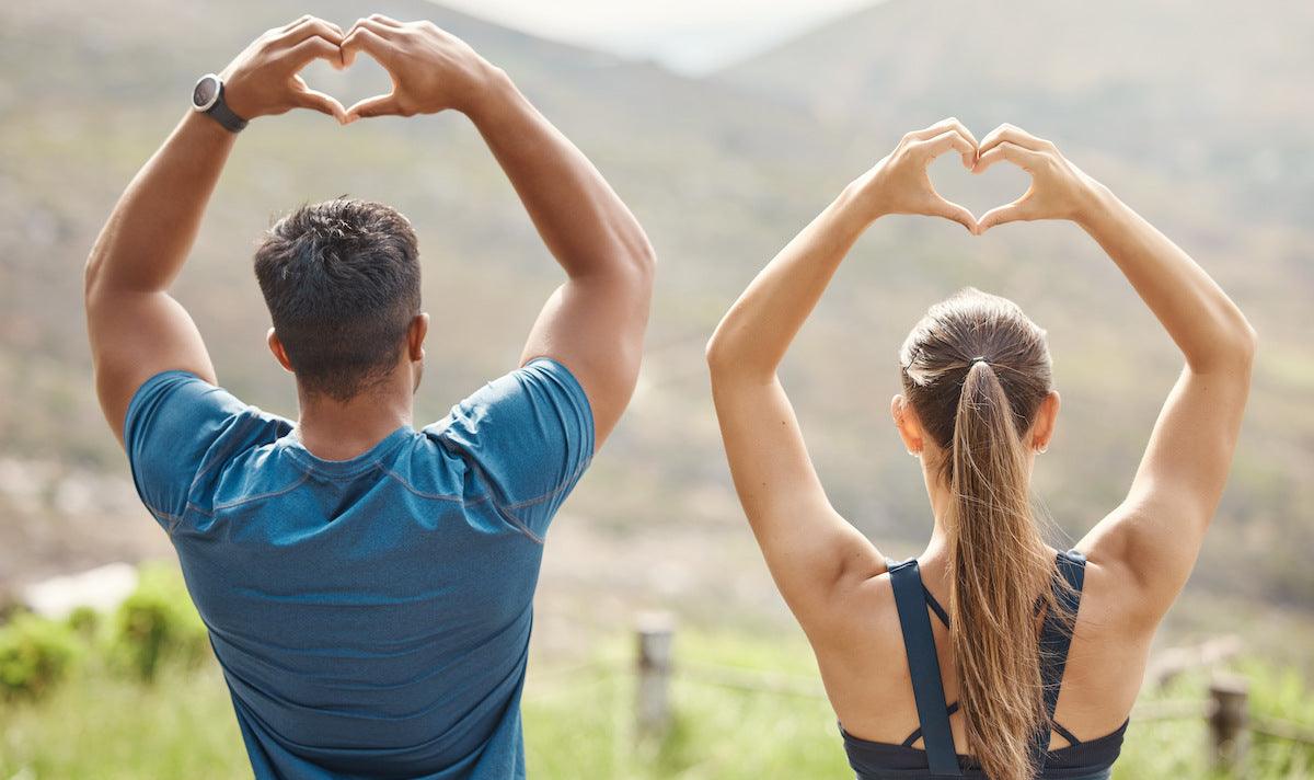 Fit young man and woman outdoors forming heart shapes with their hands above their heads, symbolizing heart health and active lifestyle.