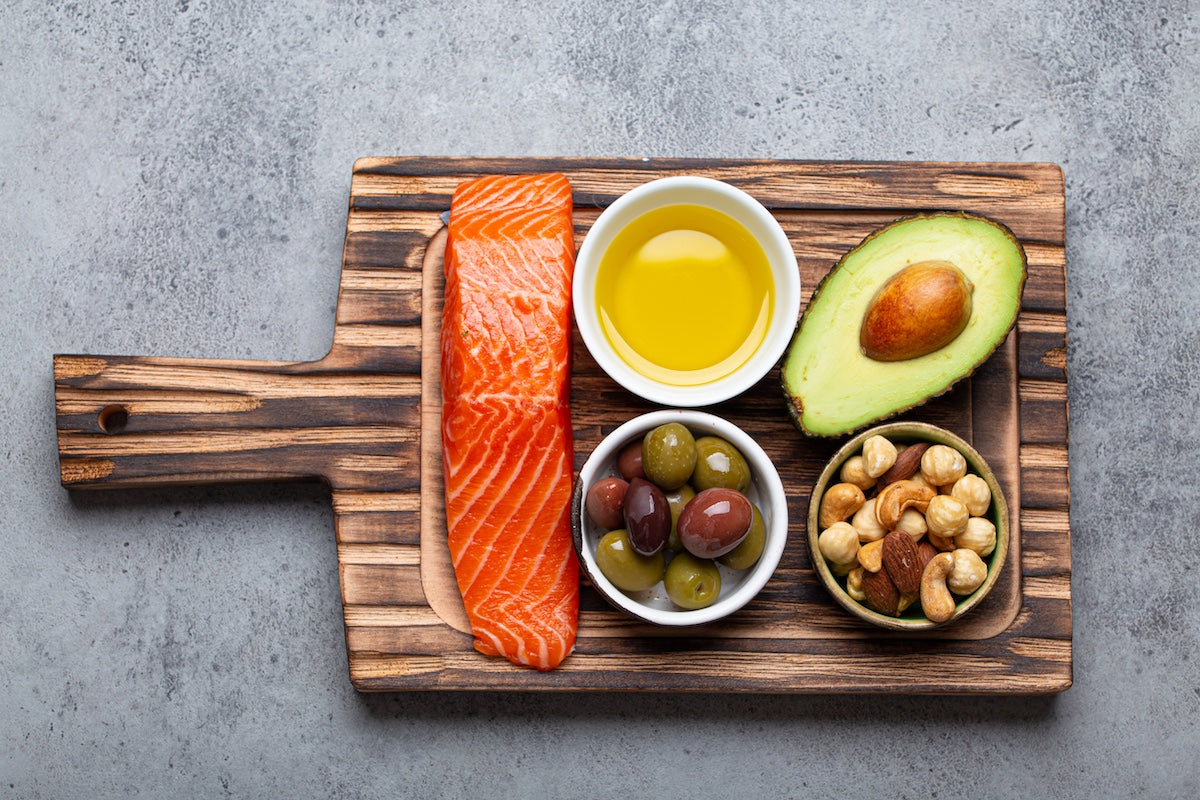 Fresh salmon fillet with olive oil, avocado, mixed olives, and nuts on a wooden board, showing natural food sources of omega-3 fatty acids and healthy unsaturated fats.