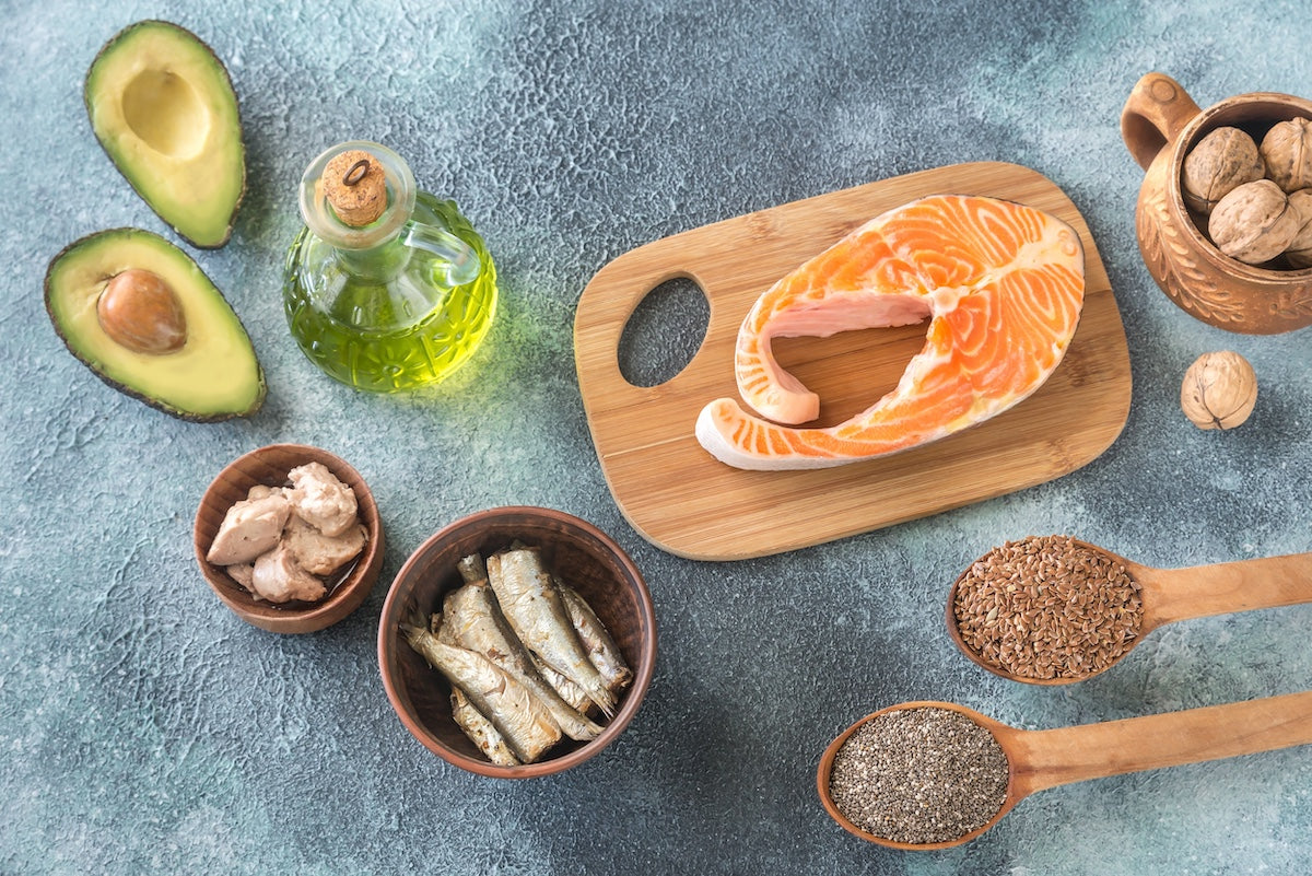 A variety of omega-3 rich foods including salmon, sardines, flaxseeds, chia seeds, avocado, walnuts, canned fish, and a bottle of olive oil arranged on a textured surface, promoting heart and blood pressure health.