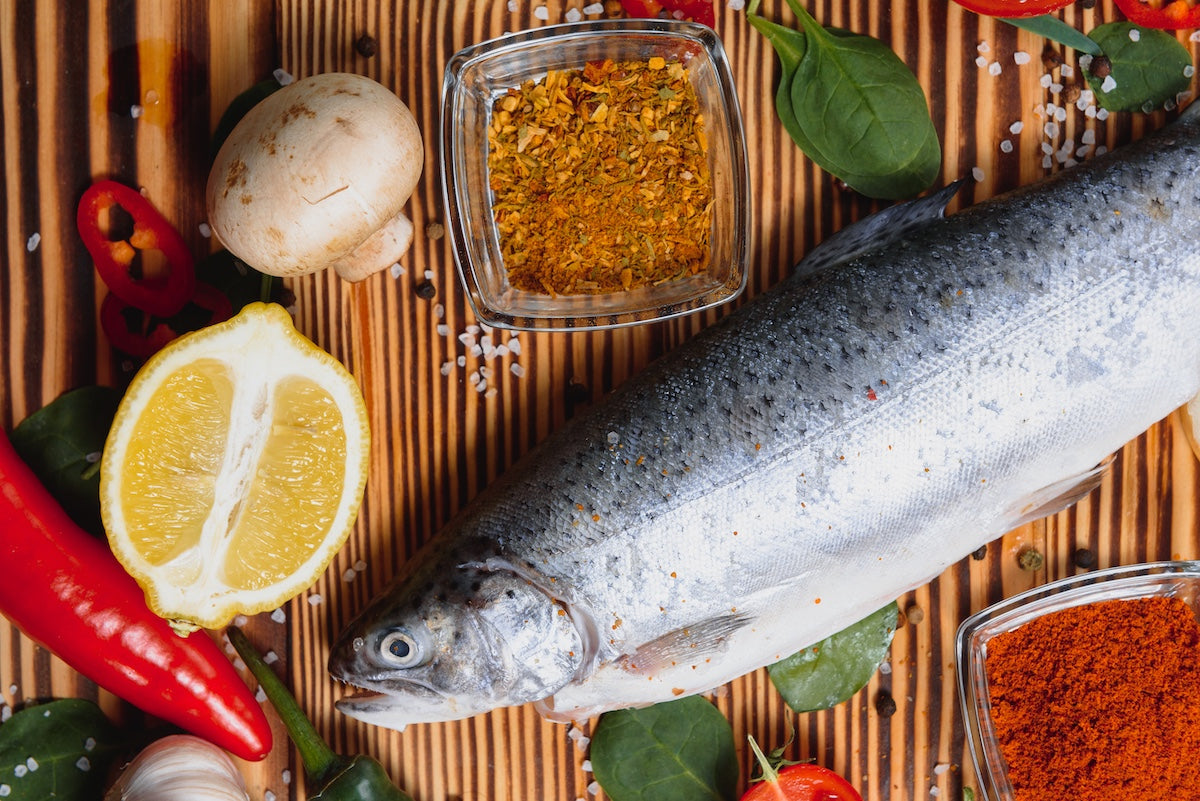 Fresh whole rainbow trout surrounded by lemon, mushroom, red chili, leafy greens, garlic, and spices on a wooden surface, illustrating a nutritious and flavorful preparation rich in omega-3 fatty acids.