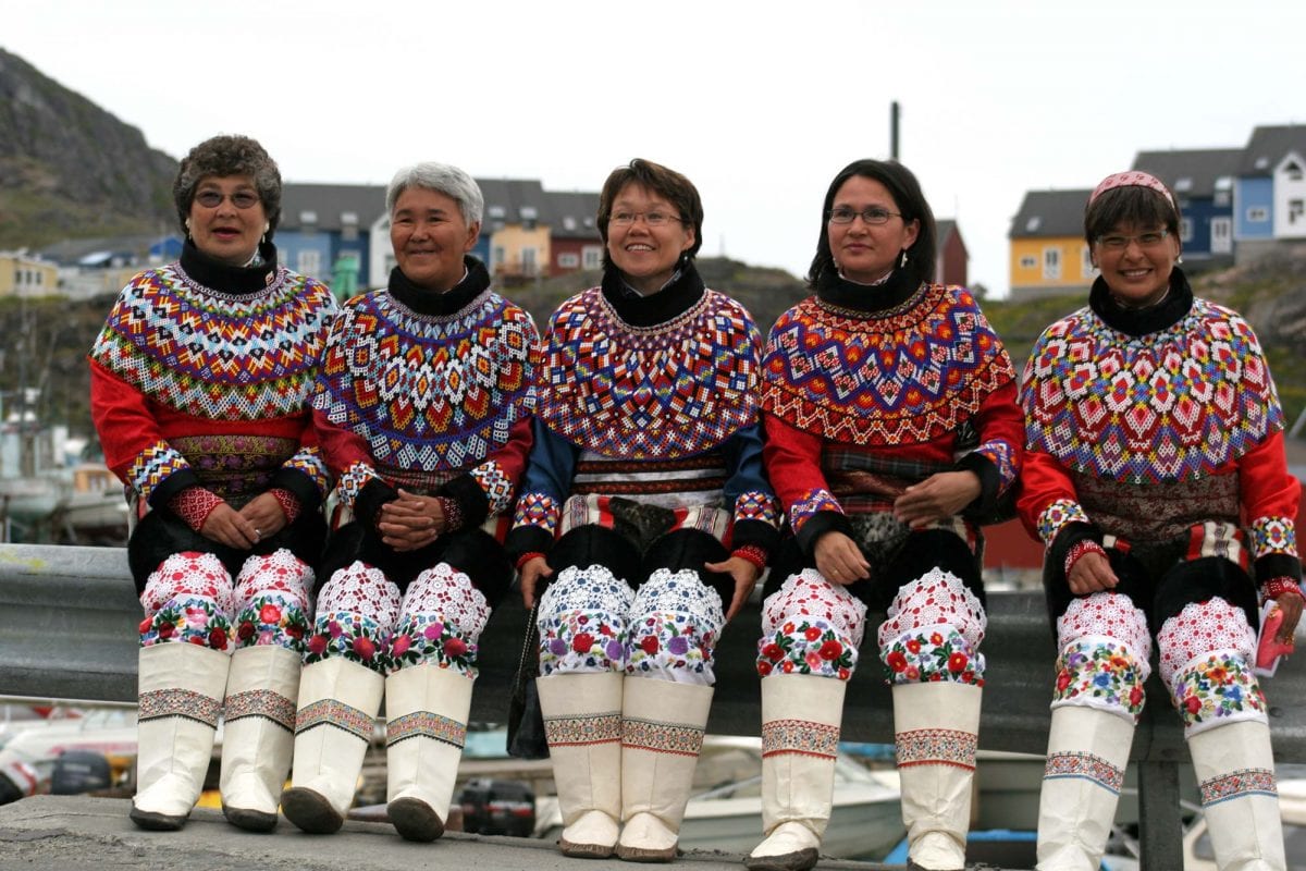 Five Greenlandic Inuit women wearing traditional Greenland national costumes with colorful beadwork and embroidery, sitting together and smiling in an outdoor setting with houses in the background.