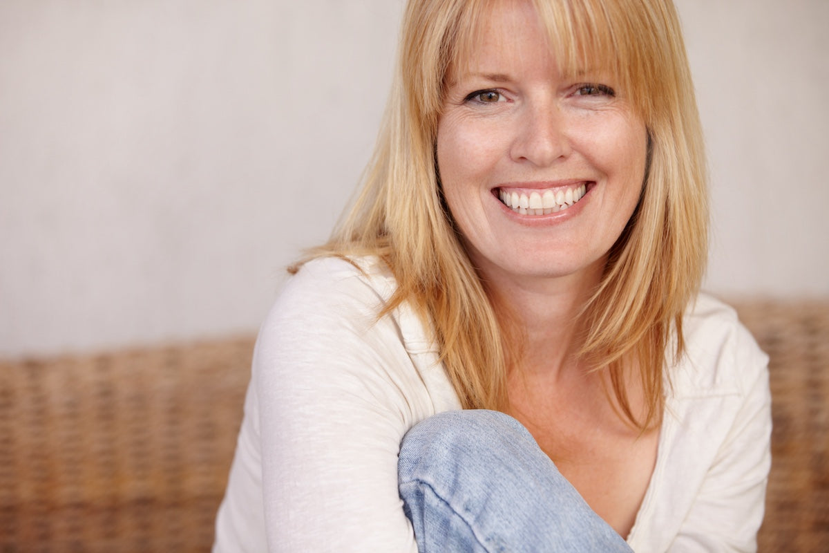 Smiling middle-aged woman with blonde hair sitting comfortably, representing healthy skin and confidence often associated with collagen supplementation.