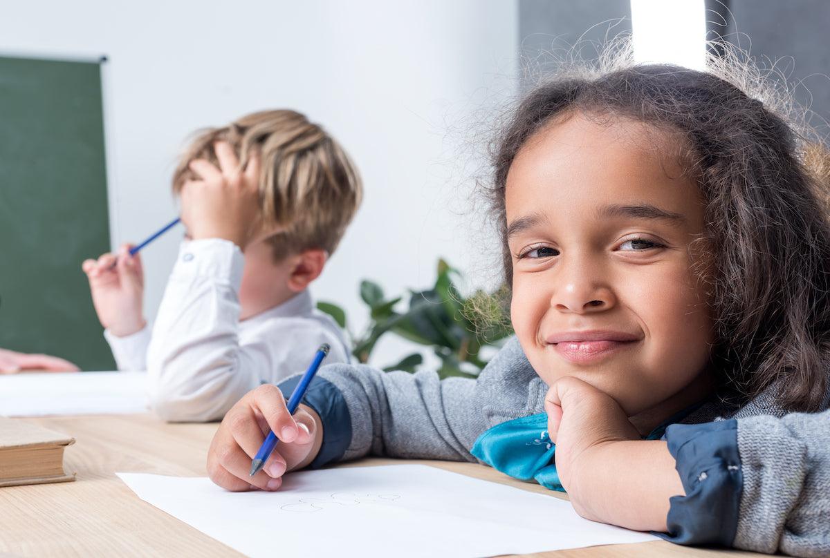Smiling young girl holding a pencil and resting her chin on her hand while drawing on paper at a classroom desk, with another child in the background appearing focused on their work.