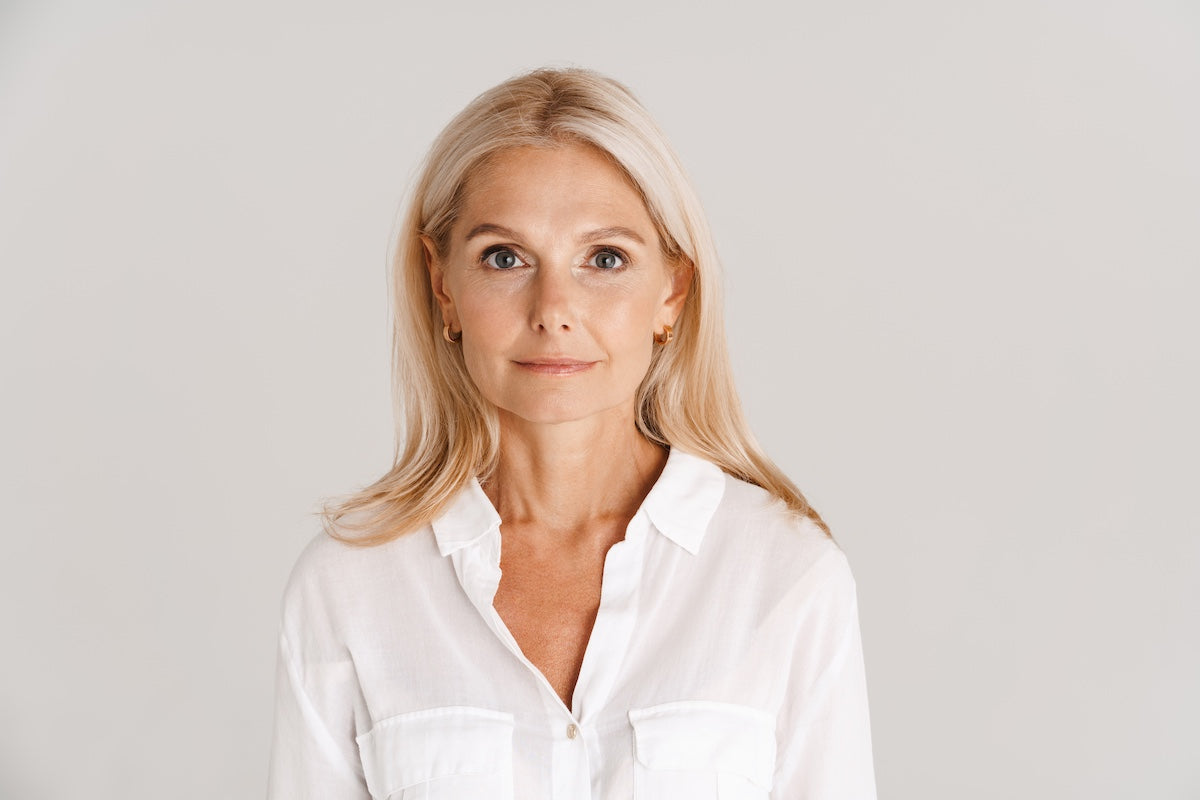 A mature blonde woman with long hair wearing a white shirt, standing against a neutral background and looking confidently at the camera.