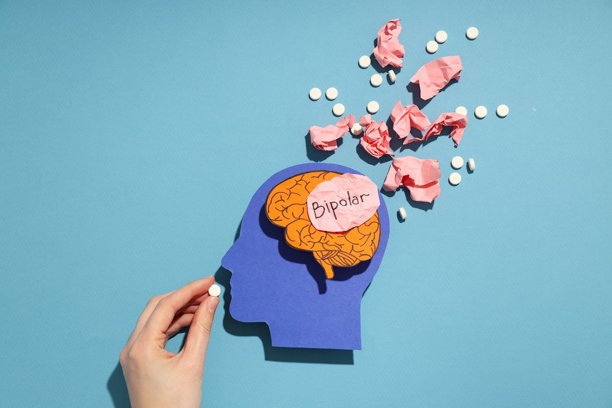 Illustration representing bipolar disorder with a cutout of a human head showing a brain labeled "Bipolar," surrounded by crumpled pink paper and scattered white pills, symbolizing mental health challenges and medication treatment.