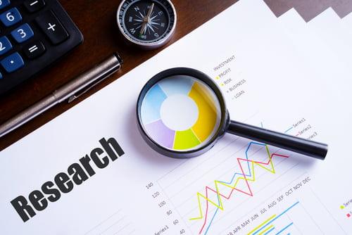 Close-up of a desk with research documents, a magnifying glass over a colorful pie chart, a compass, a pen, and a calculator, symbolizing data analysis and academic or scientific investigation.