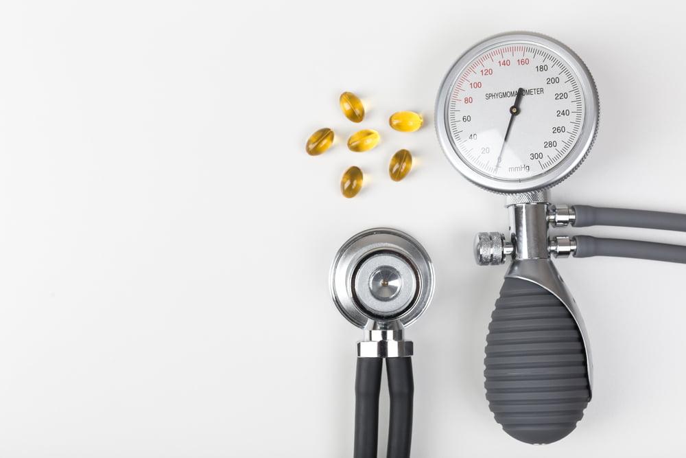 Blood pressure monitor (sphygmomanometer) and stethoscope placed on a white background next to several yellow omega-3 capsules, symbolizing the connection between heart health, blood pressure, and omega-3 supplements.