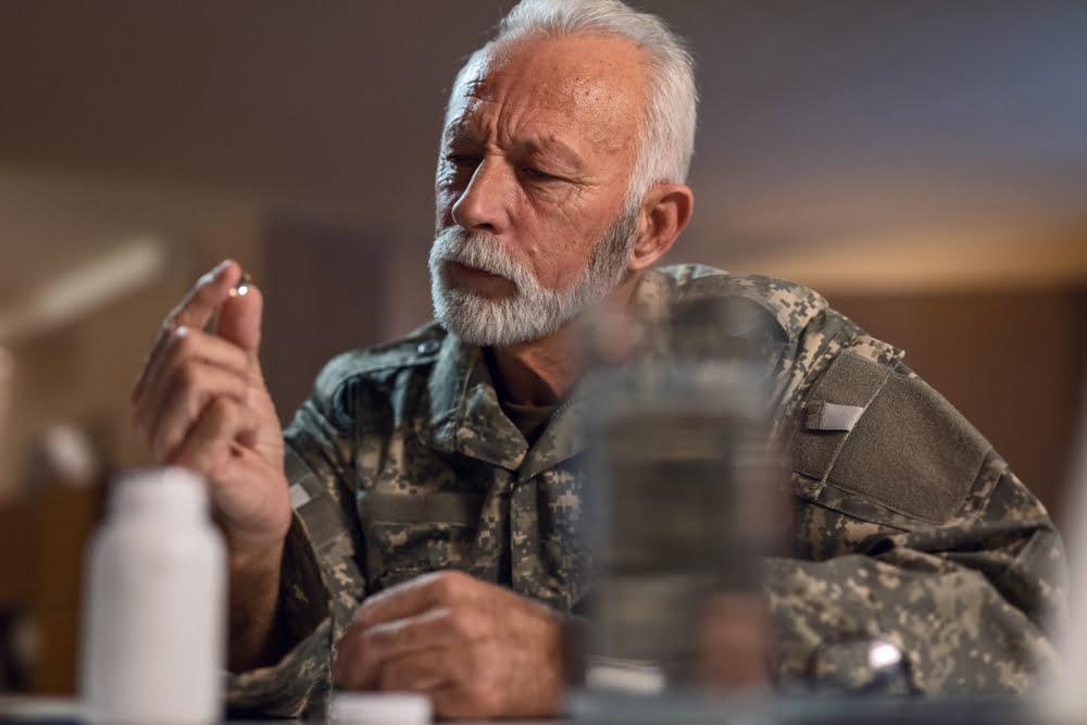 Elderly man in military uniform examining a supplement capsule while seated at a table with medicine bottles, symbolizing health awareness and supplement use among veterans or older adults.