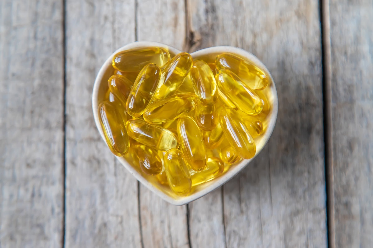 Heart-shaped bowl filled with golden omega-3 fish oil capsules on a rustic wooden surface, symbolizing heart health and nutritional supplements.