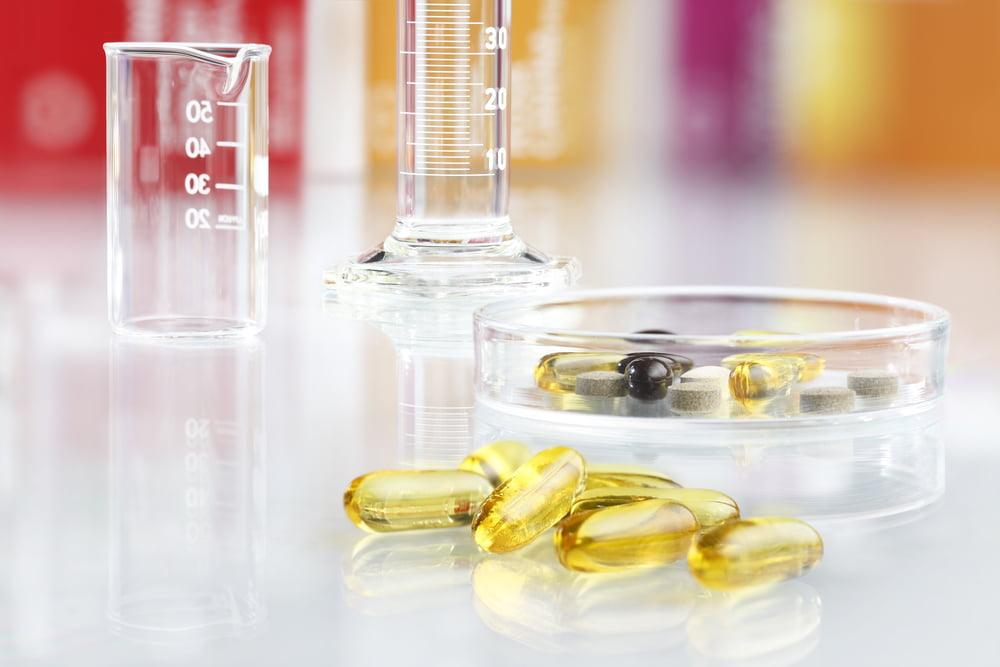 A scientific lab setting showing transparent capsules of fish oil on a glossy surface, alongside a petri dish containing various supplements, a small beaker, and a graduated cylinder.