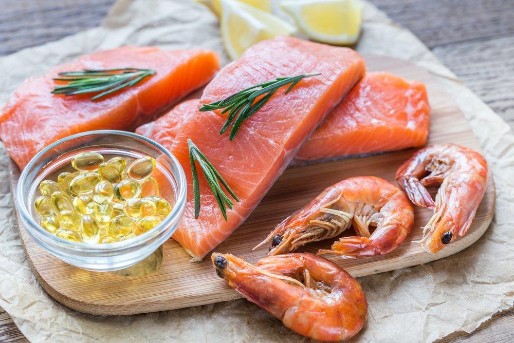 A platter of salmon, herbs, shrimps and omega-3 capsules on a wooden cutting board with lemons next to it. 