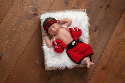 A newborn baby dressed in a red and black crocheted boxer outfit, with tiny boxing gloves, peacefully sleeping on a fluffy white cushion inside a wooden box on a wooden floor.