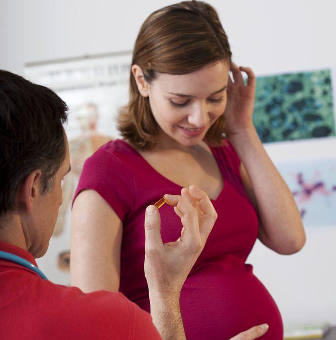 A pregnant woman in a red shirt smiles as a healthcare professional offers her a softgel capsule, possibly a prenatal omega-3 supplement, during a consultation in a medical office.