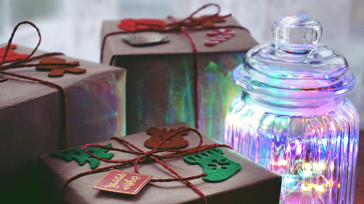 Christmas presents wrapped in brown paper with festive decorations beside a glass jar glowing with colorful holiday lights