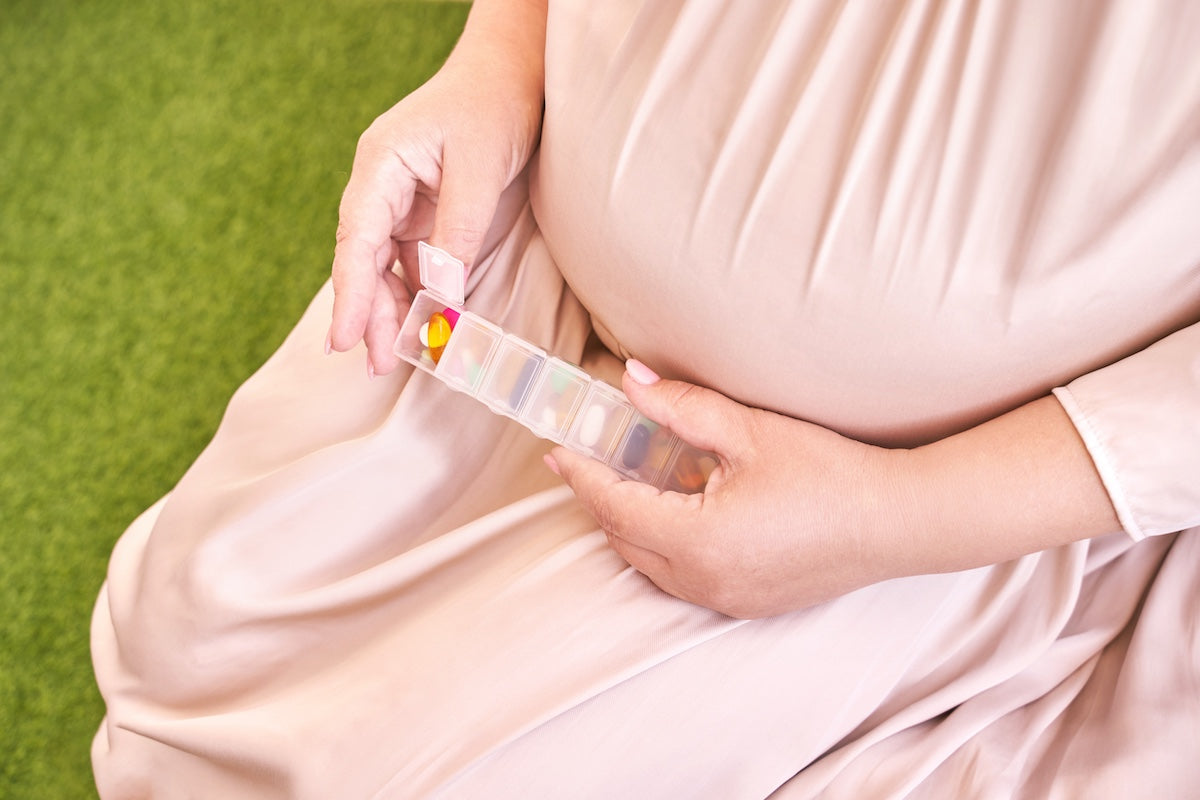 Pregnant woman in a pink dress sitting on the floor and holding a weekly pill organizer filled with multicolored prenatal vitamins and supplements.