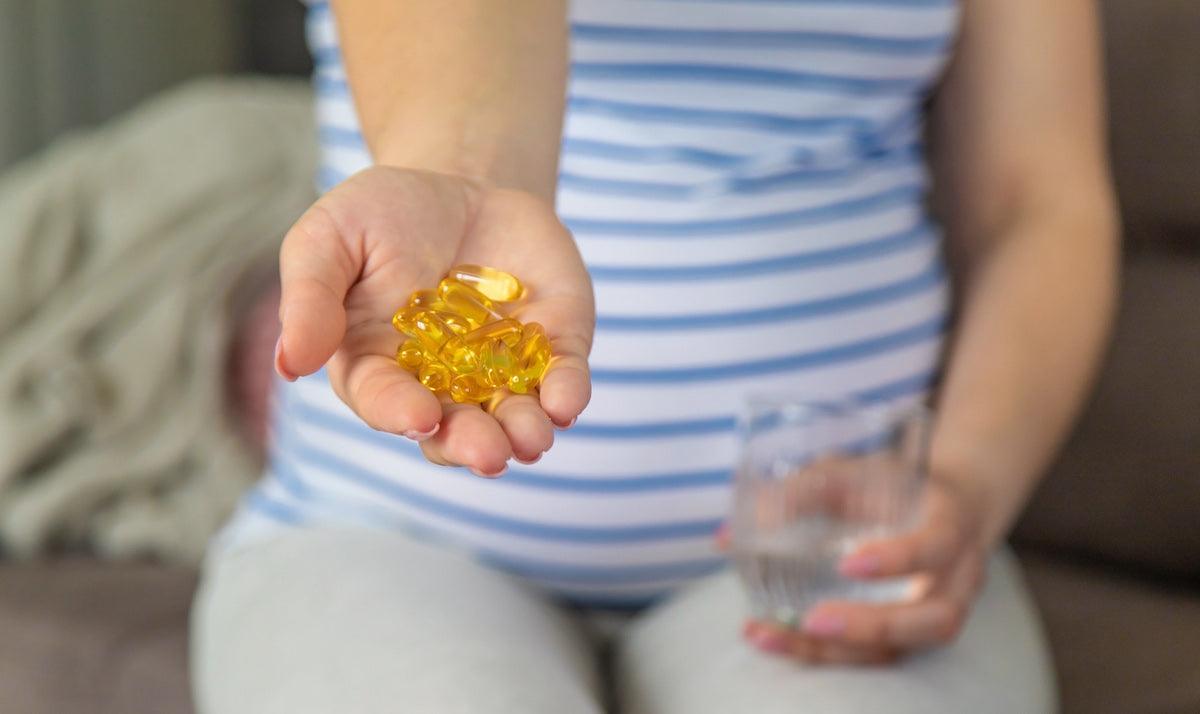 Pregnant woman in a striped shirt holding yellow omega-3 fish oil capsules in one hand and a glass of water in the other, highlighting the importance of DHA supplements during pregnancy.