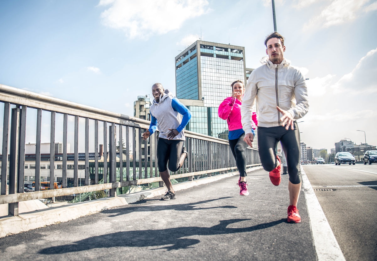 Three athletic individuals running outdoors on an urban bridge, dressed in sporty attire with buildings and city traffic in the background, representing active lifestyle and fitness.
