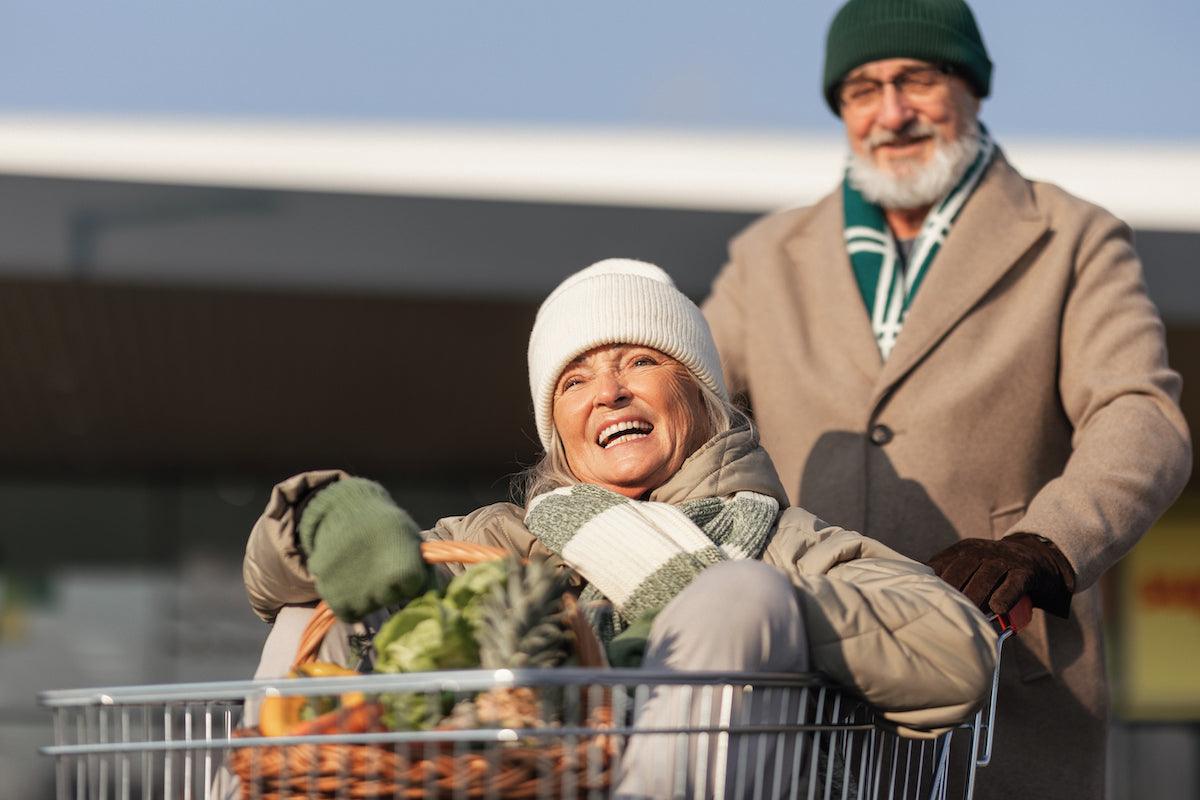 Smiling senior woman sitting in a shopping cart filled with groceries, being pushed by her partner outside a grocery store on a sunny day.