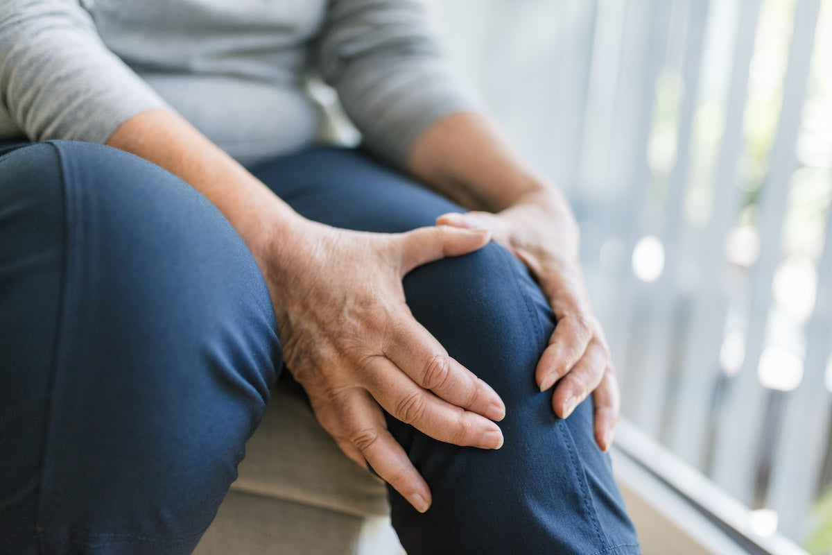 Senior woman gently holding her knee with both hands, indicating joint pain or discomfort, seated indoors near a window with natural light.