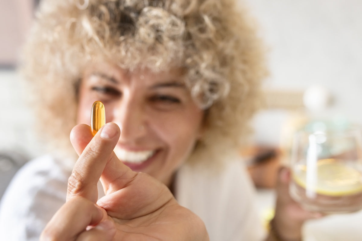Smiling woman holding a golden omega-3 supplement capsule between her fingers, with a glass of water in the background, promoting daily health and wellness.
