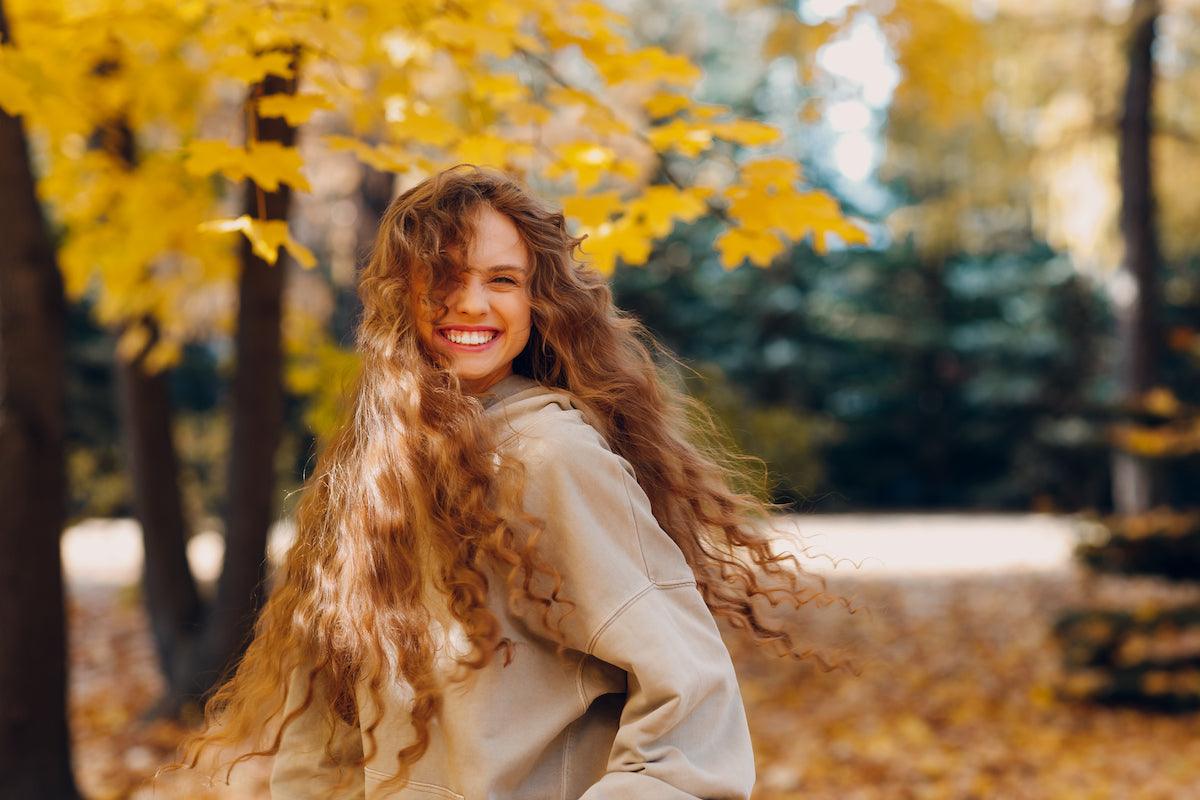 Smiling young woman with long, curly hair wearing a beige hoodie, standing outdoors in a park with yellow autumn leaves in the background.