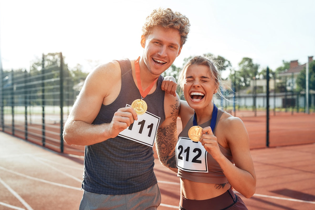 Smiling male and female track athletes proudly holding gold medals while standing on an outdoor running track, wearing race bibs and athletic gear.