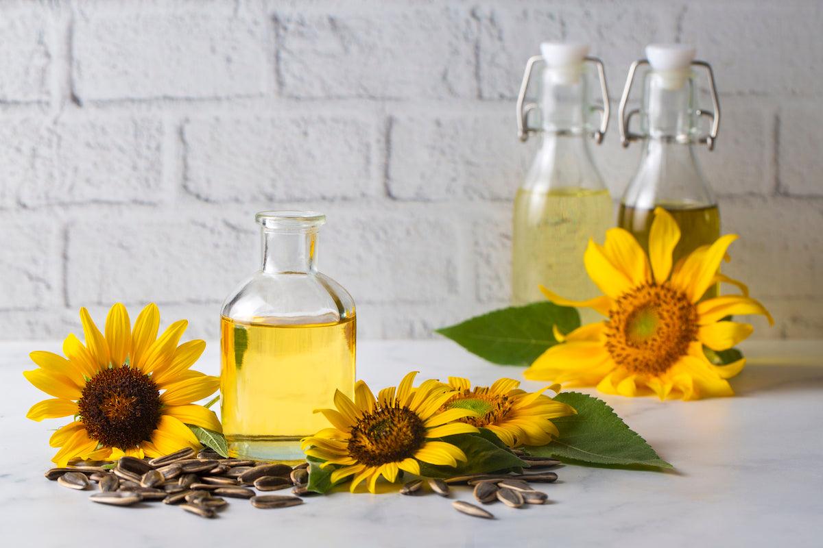 Glass bottles of sunflower oil accompanied by vibrant sunflower blossoms and scattered sunflower seeds, set against a light brick background.