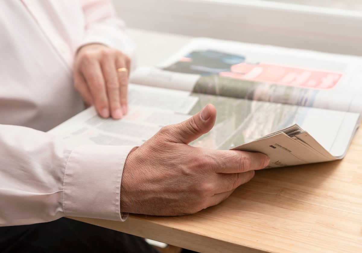 Close-up of an older man’s hands holding and reading a newspaper at a wooden table, dressed in a light pink shirt, suggesting a moment of relaxed focus or staying informed.