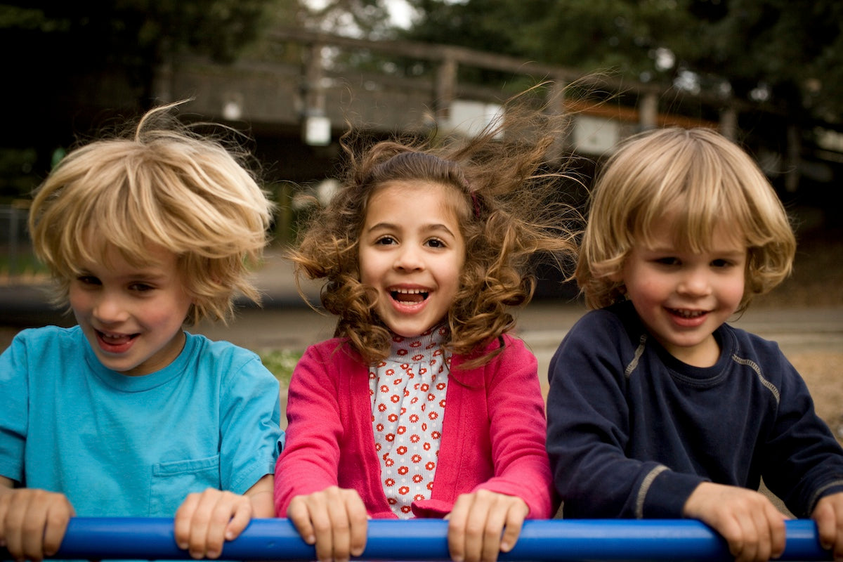 Three smiling children playing outdoors, holding onto a blue bar with windblown hair, enjoying a fun moment at the playground.