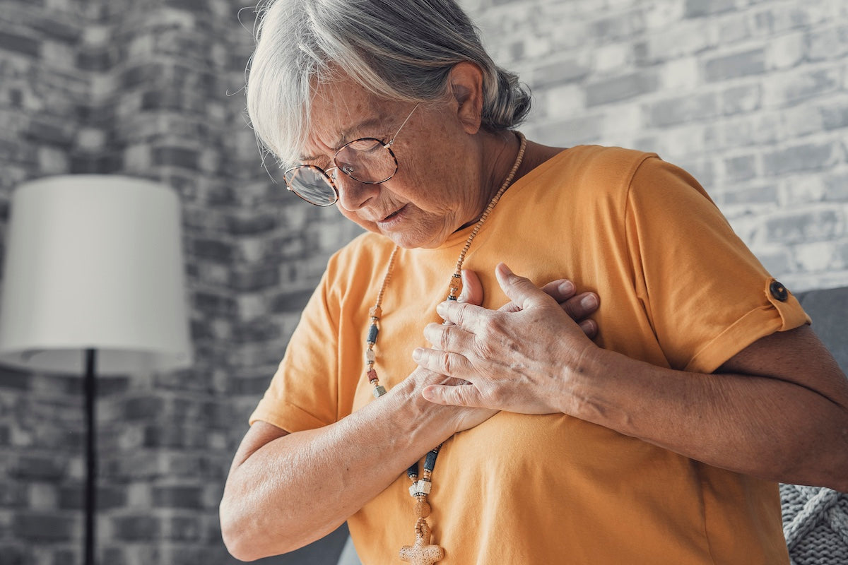 Elderly woman wearing glasses and an orange shirt clutching her chest in discomfort, possibly experiencing chest pain or early symptoms of a heart problem.