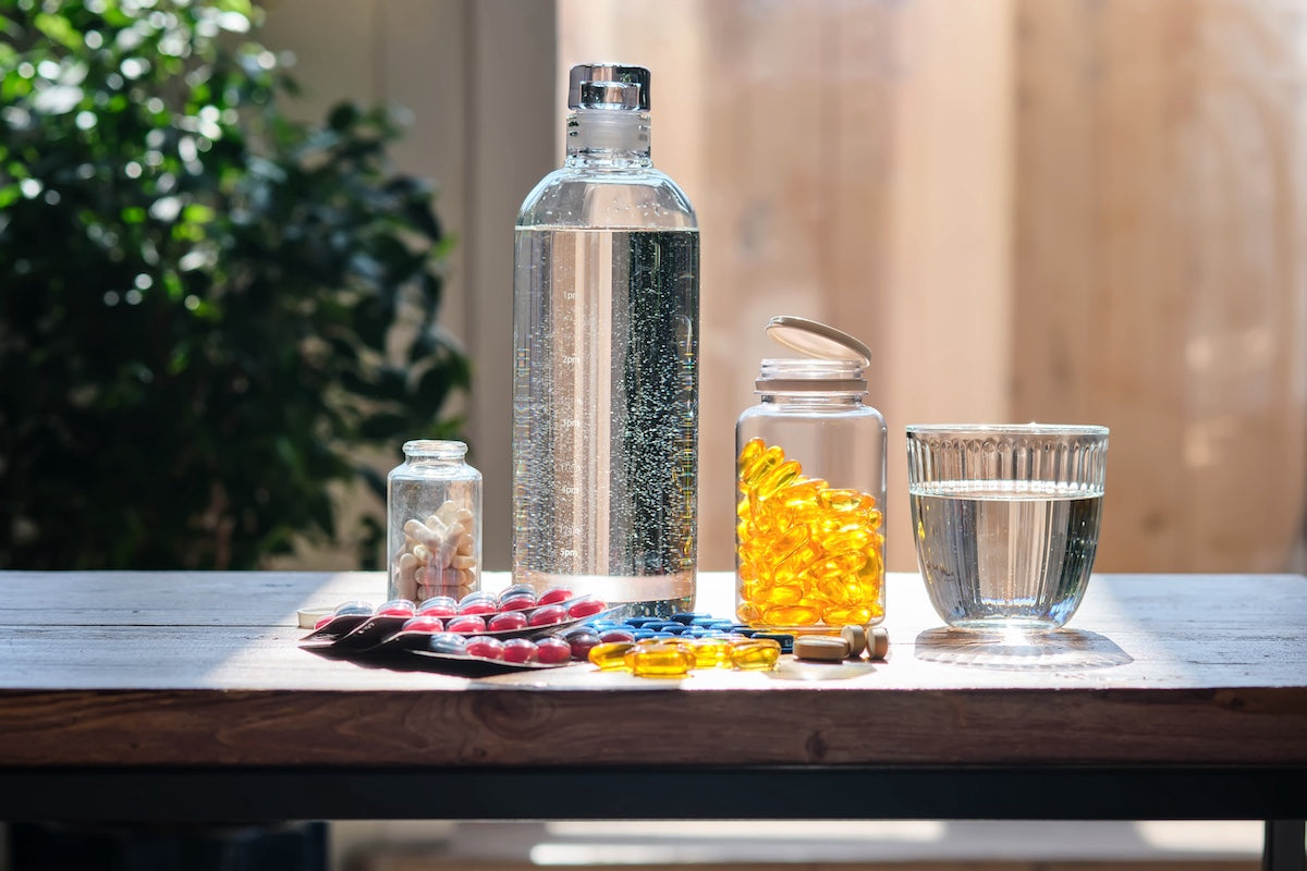 Various dietary supplements, including omega-3 fish oil capsules, multivitamins, and medications, arranged on a wooden table next to a glass and bottle of water, with natural sunlight and greenery in the background.