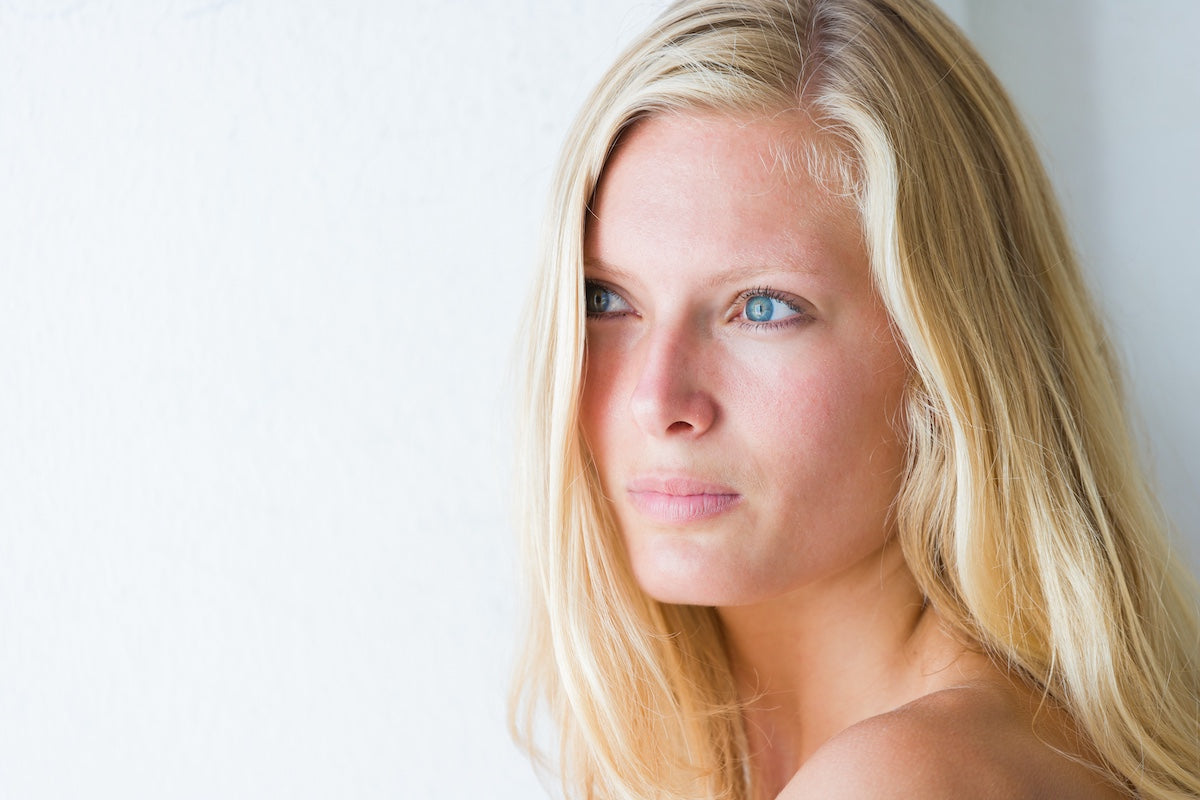 A fair-skinned, blonde-haired, blue-eyed young woman stands in front of a white background; representing the themes of healthy skin, beauty, and collagen supplementation.