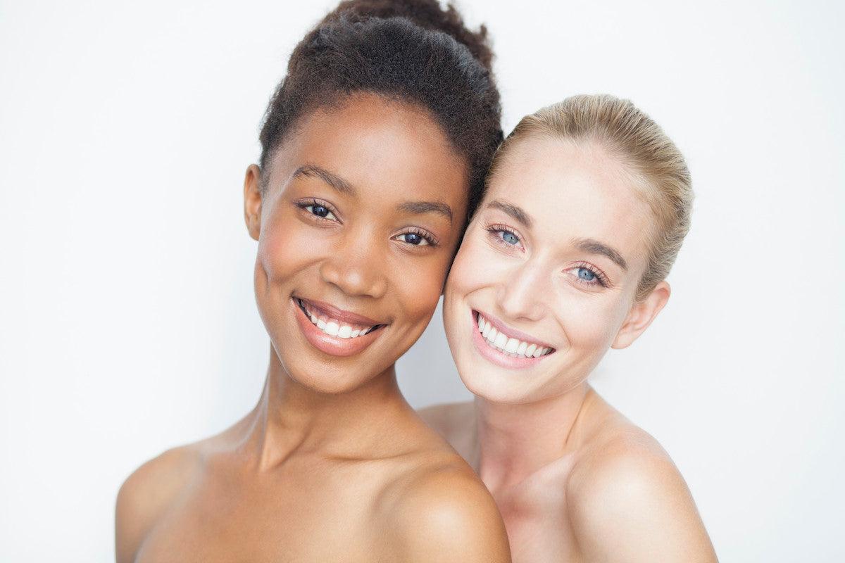 Two smiling women with radiant, healthy skin and bare shoulders standing close together against a light background.