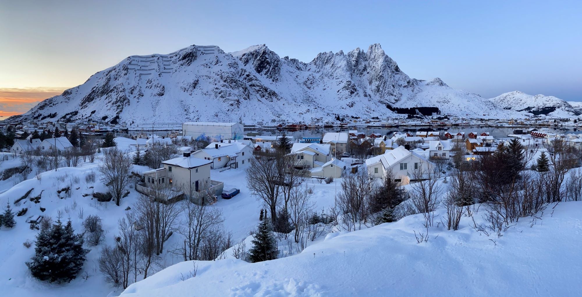 Snow-covered village nestled at the base of rugged mountains in Lofoten, Norway, during a serene winter sunset with pastel skies and scattered trees in the foreground.