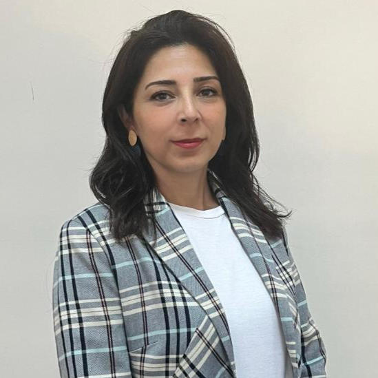 Derya Karakoyun with shoulder-length dark hair, wearing a plaid blazer over a white shirt and gold earrings, posing against a plain beige background.