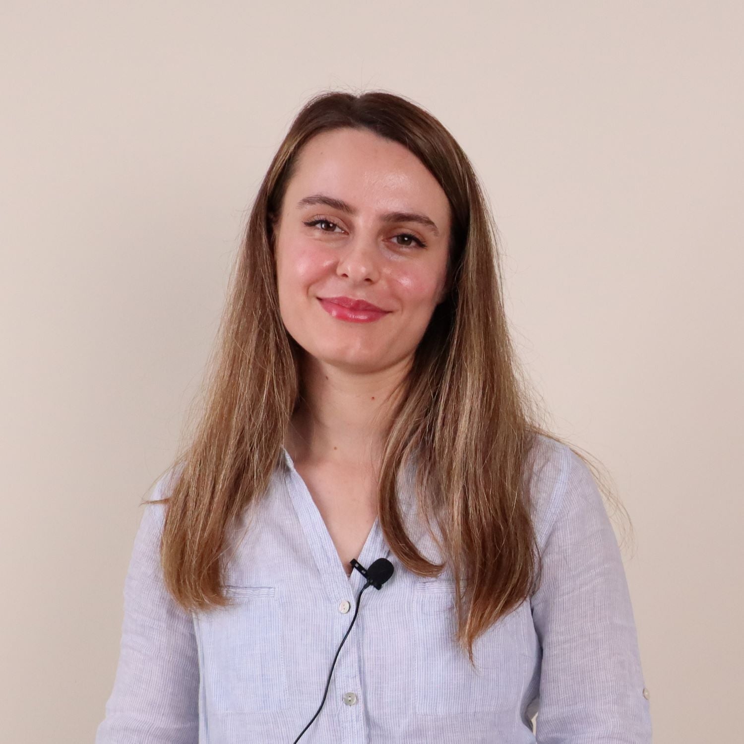 Smiling Fatma Beğboğa with long, light brown hair, wearing a light blue button-up shirt and a clip-on microphone, posing against a beige background.