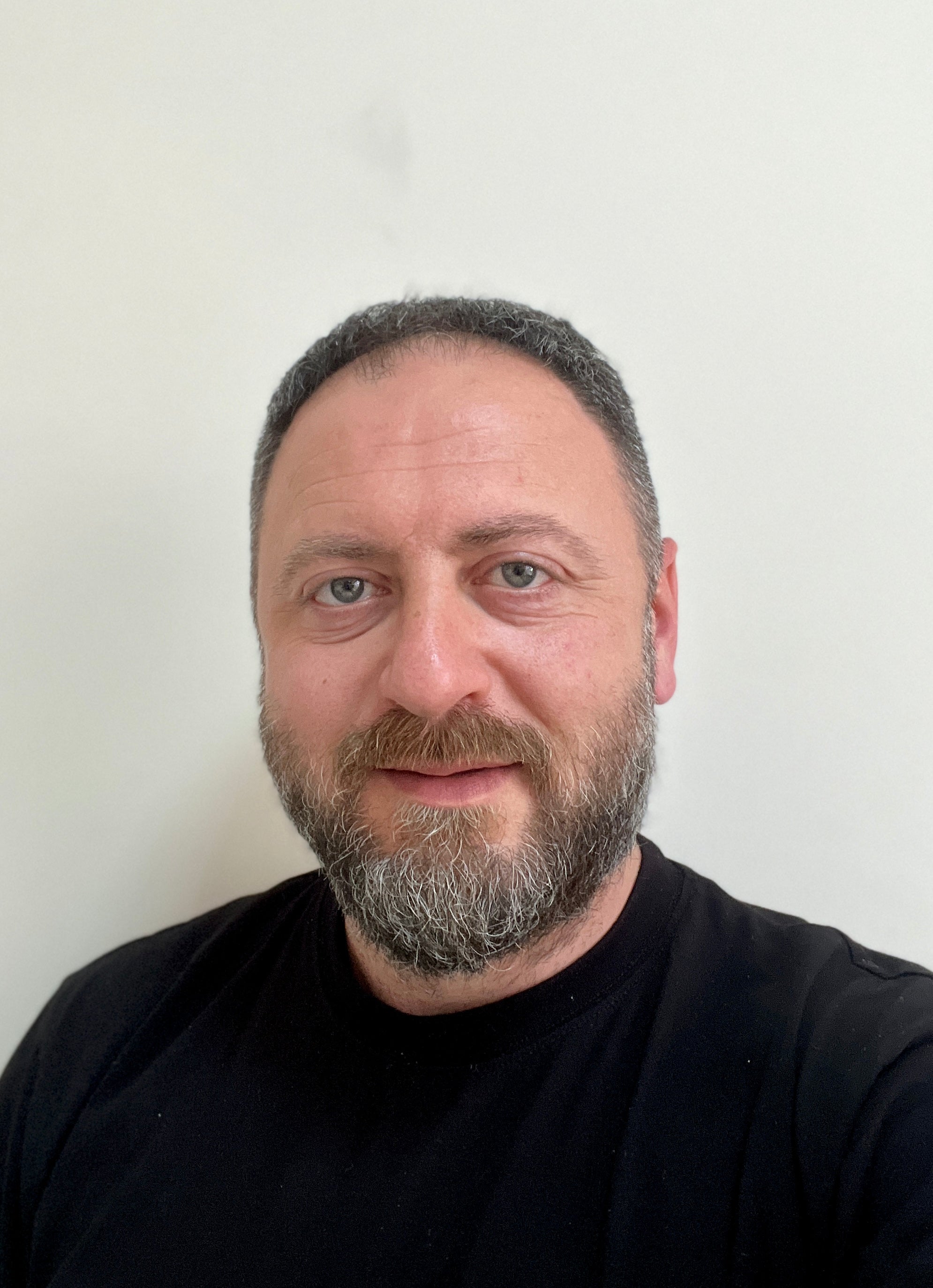Smiling İldem Kulli with short, dark hair, a well-groomed beard, and light skin, wearing a black T-shirt, posing against a plain white background.