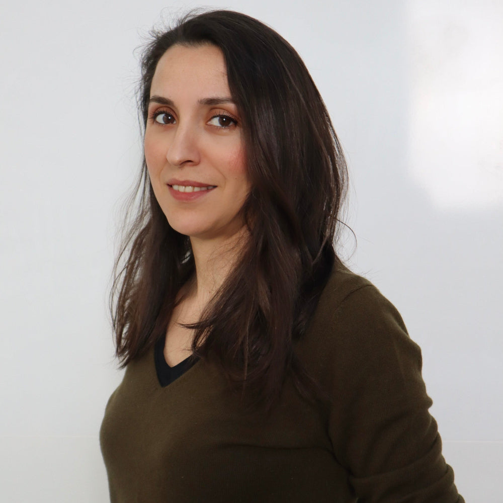 Smiling Meryem Kulli with long, dark brown hair, wearing an olive green sweater with a black V-neck detail, posing against a white background.