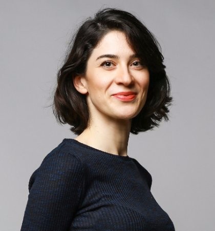 Smiling Mezago Kuyucu with short, wavy dark hair, wearing a navy blue textured sweater, posing against a neutral gray background.