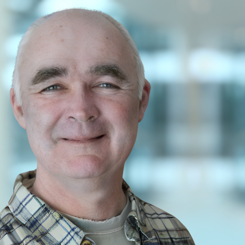 Smiling Per Benjaminsen with light skin, bald head, and thick dark eyebrows, wearing a plaid shirt and a beige undershirt, standing in a bright indoor setting with blurred background.