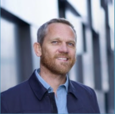 Smiling Børge Bjelland, wearing a navy blue jacket over a light blue shirt, standing outdoors with a modern glass building in the background.