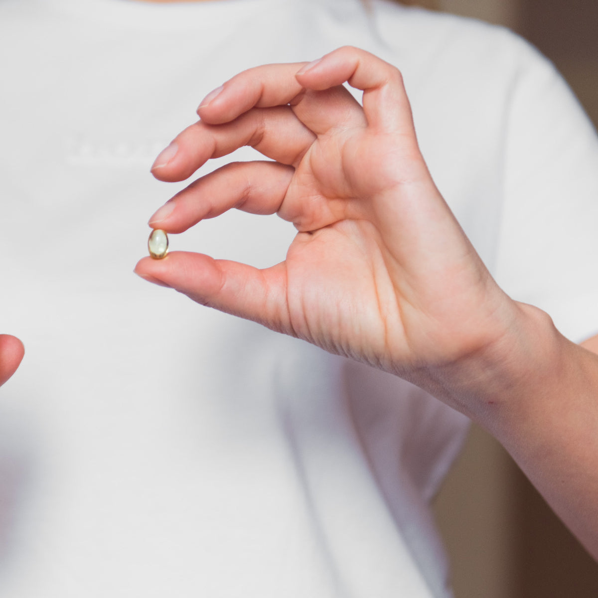Close-up of a person in a white shirt holding a single fish oil capsule between their fingers.