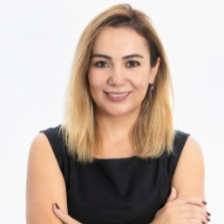 Smiling Özge Olsen with shoulder-length blonde hair, wearing a sleeveless black dress, posing with her arms crossed against a plain white background.